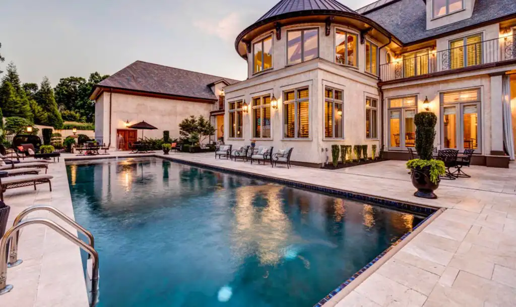 Large stone home at dusk with a long lit pool and travertine deck