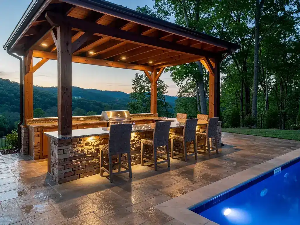 Stone outdoor kitchen with a built-in grill and bar seating beside a lit pool at dusk
